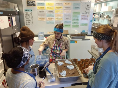 Four people in a kitchen preparing desserts, with one mixing ingredients and others filling cups. Colorful aprons and headbands worn.