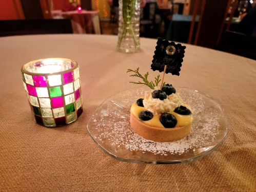 A dessert on a glass plate with blueberries and cream, next to a colorful candle on a table.