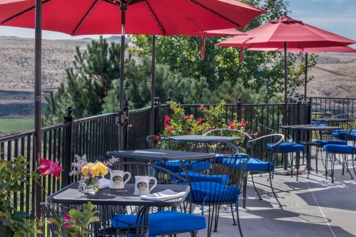 Outdoor dining area with blue chairs, tables, and red umbrellas, surrounded by greenery and scenic hills in the background.