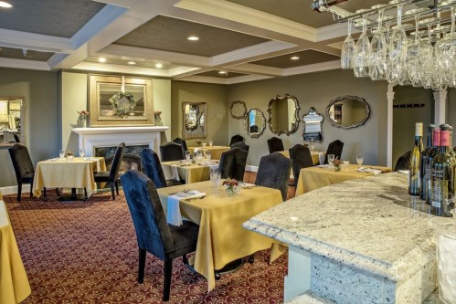 Elegant restaurant interior with tables set in yellow linens, black chairs, and a cozy fireplace. Mirrors adorn the walls.