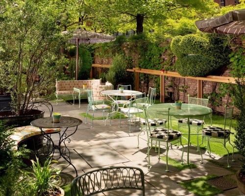 A serene outdoor patio with green chairs and tables, surrounded by lush plants and shaded by umbrellas.