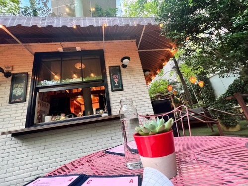 A cozy outdoor dining area with a pink table, a succulent plant, and a view of a charming restaurant with string lights.