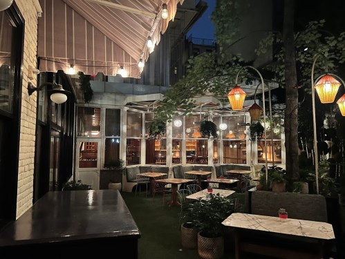 A cozy outdoor restaurant at night, featuring tables, plants, and warm lantern lighting under a striped awning.