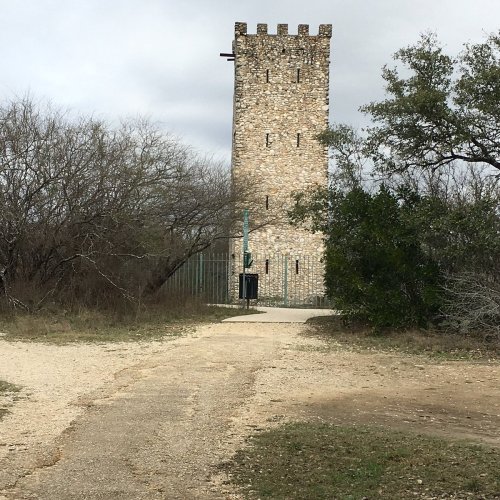 A tall stone tower stands at the end of a gravel path, surrounded by sparse trees and cloudy skies.