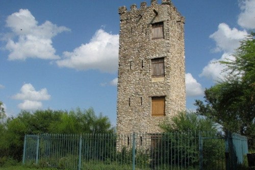 A tall, stone tower with boarded windows, surrounded by greenery and a metal fence under a blue sky with clouds.