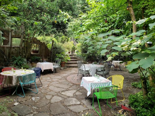 A serene outdoor dining area with colorful tables and chairs surrounded by lush greenery and string lights.