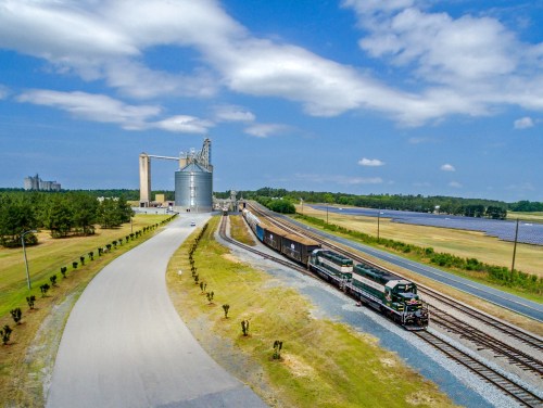 A train travels along tracks near grain silos, with a clear blue sky and fields in the background.