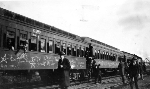 Black and white photo of a train with graffiti, people boarding, and standing on the tracks in a historical setting.