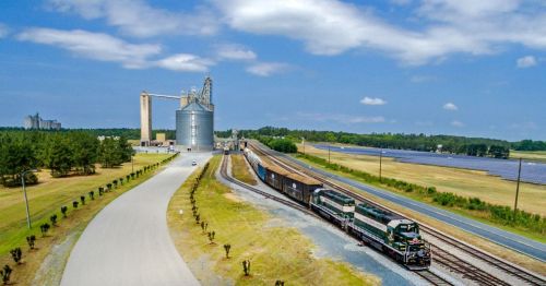 A train passes by a grain silo and fields under a blue sky, with a road and solar panels in the background.