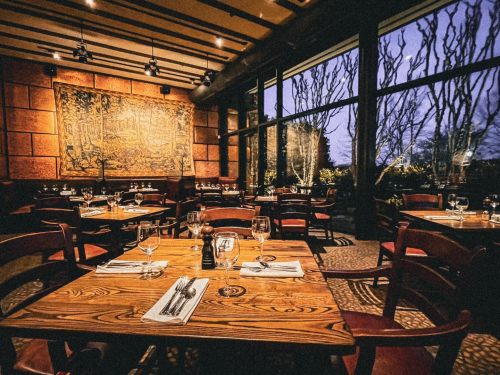 Cozy restaurant interior with wooden tables, chairs, and large windows showcasing a twilight view outside.