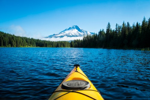 A yellow kayak on a calm lake, with a snow-capped mountain and dense forest in the background under a clear blue sky.