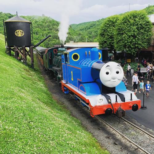 A colorful cartoon train with a smiling face, surrounded by greenery and people, at a scenic railway station.
