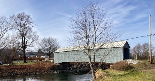 A blue covered bridge spans a calm river, surrounded by bare trees and a clear sky.