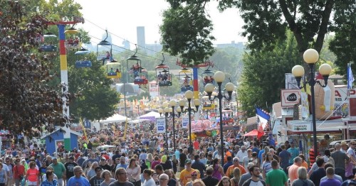 A bustling fairground filled with crowds, colorful booths, and a scenic view of the skyline in the background.