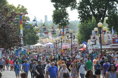 A bustling fairground scene with crowds of people, colorful stalls, and overhead gondolas in a sunny outdoor setting.