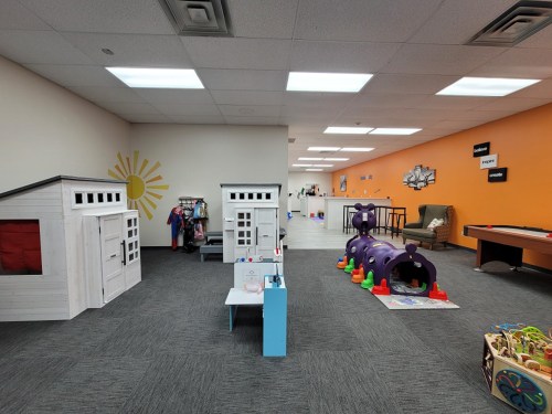 Bright indoor play area featuring playhouses, a colorful tunnel, and various toys on the floor.