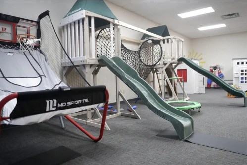 Indoor play area featuring a basketball hoop, climbing structure, slide, and various sports equipment.