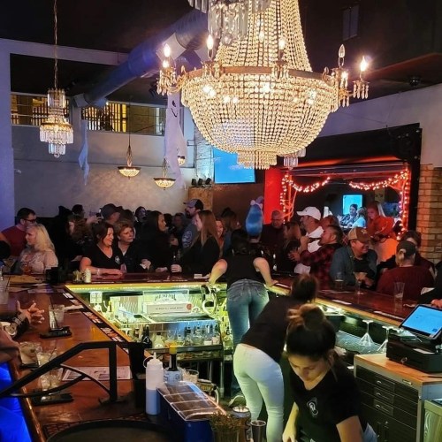 A lively bar scene with a chandelier, crowded patrons, and bartenders serving drinks. Warm, festive atmosphere.