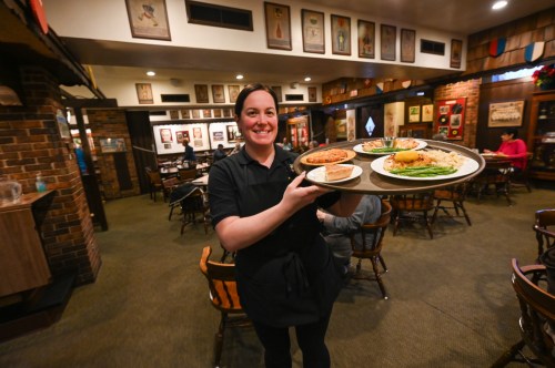A smiling waitress carries a tray with food in a cozy restaurant setting, with diners in the background.