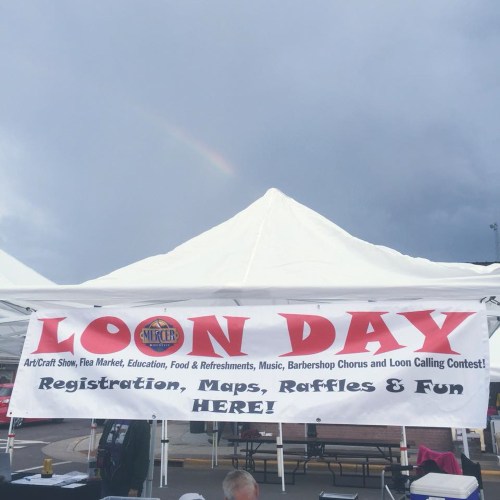 A banner for "Loon Day" event featuring activities like crafts, food, and a contest, under a cloudy sky with a rainbow.