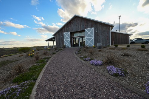 A rustic building with white doors, surrounded by a gravel path and blooming flowers under a cloudy sky.