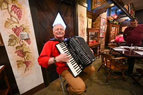 An elderly man in a red shirt plays the accordion while seated in a cozy restaurant with rustic decor.