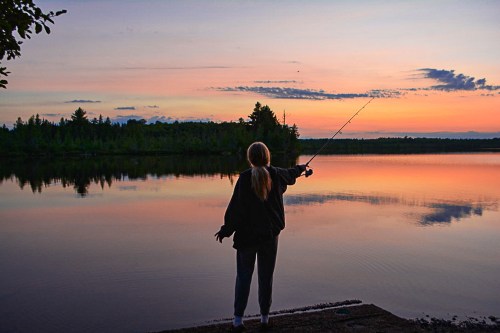 A person fishing at sunset by a calm lake, surrounded by trees and reflecting colorful skies.