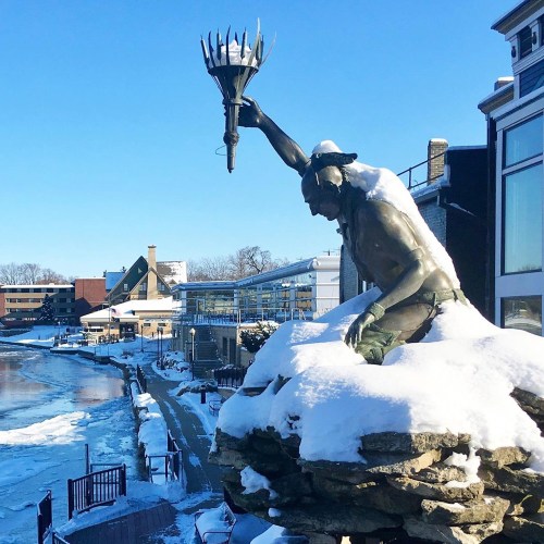 A snow-covered statue of a Native American holding a torch, overlooking a frozen river and buildings in winter.