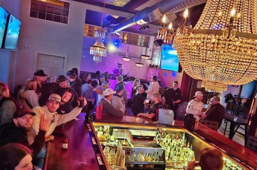 A lively bar scene with a crowd socializing under a large chandelier, colorful lights, and a well-stocked bar.