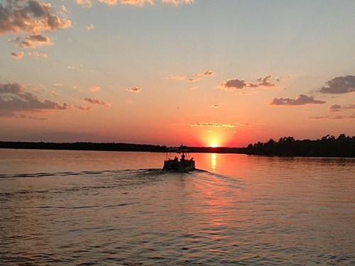 A boat glides across a calm lake at sunset, with vibrant orange and pink hues in the sky and clouds.
