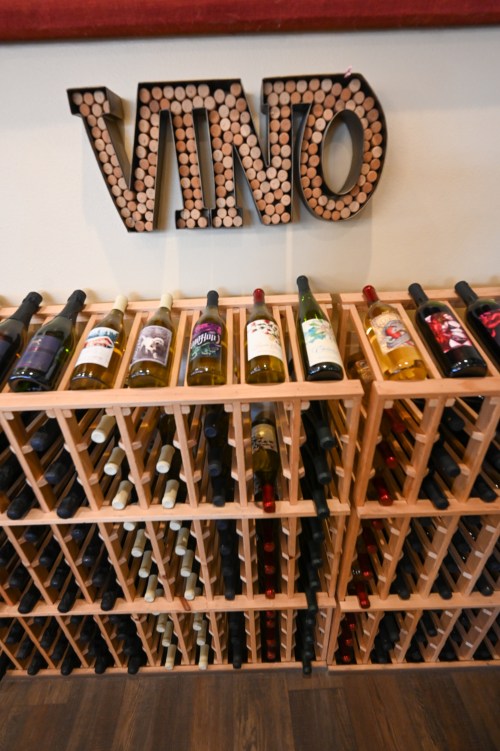 A wooden wine rack displaying various bottles of wine, with a decorative "VINO" sign made of corks above.