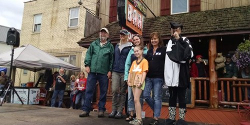 A group of six people stands on a stage in front of a bar, smiling and posing for a photo during an event.