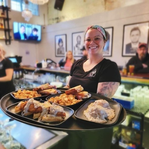 A smiling server holds a tray with multiple plates of food, including sandwiches and sides, in a lively restaurant setting.