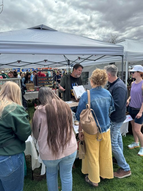 A vendor talks to a group of people at a busy outdoor market under gray skies and tents.