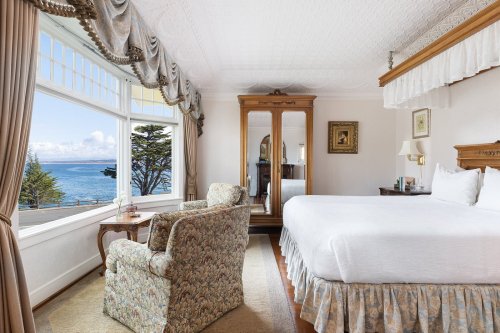 Cozy hotel room with a bed, floral armchair, and large window showcasing a scenic ocean view.