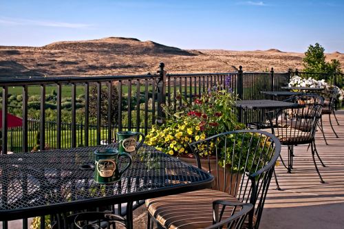 A scenic outdoor patio with black metal tables, chairs, and colorful flower pots, overlooking rolling hills.