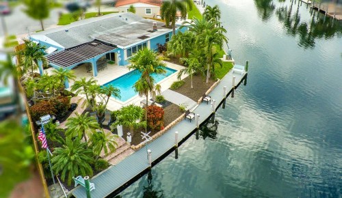 Aerial view of a waterfront home with a pool, palm trees, and a dock along a calm river.