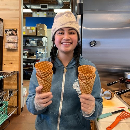 A smiling woman in a blue jacket and hat holds two ice cream cones in a kitchen setting.