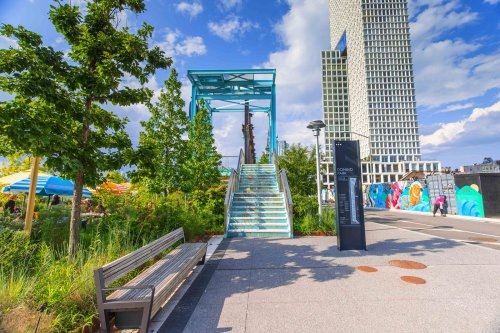 A modern urban park with greenery, a staircase, and a tall building in the background under a blue sky.