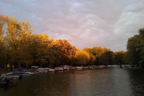 A serene river scene with boats docked along the shore, surrounded by trees displaying autumn colors under a cloudy sky.
