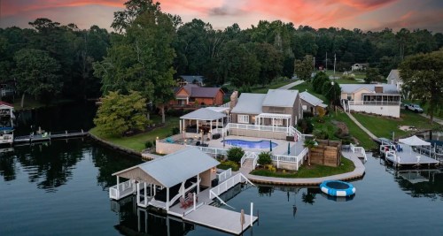 Aerial view of a lakeside home with a pool, dock, and surrounding greenery at sunset.