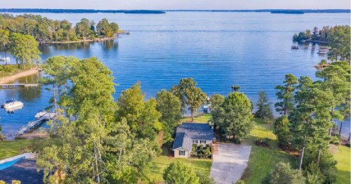Aerial view of a lakeside home surrounded by trees, with calm water and boats in the distance.