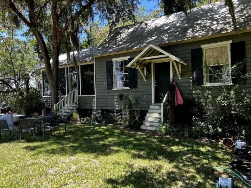 A charming green house with a porch, surrounded by trees and a grassy yard, featuring black shutters and a pink item hanging.