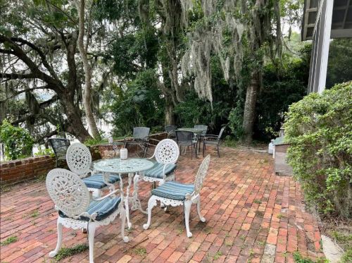 A cozy outdoor seating area with white metal furniture on a brick patio, surrounded by lush greenery and trees.