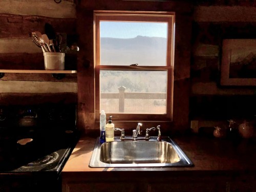 A rustic kitchen with a stainless steel sink, window view of mountains, and wooden shelves with utensils.