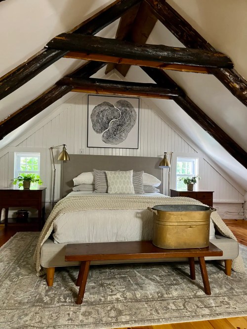 Cozy attic bedroom with a bed, wooden beams, artwork, and a vintage metal tub on a bench at the foot of the bed.