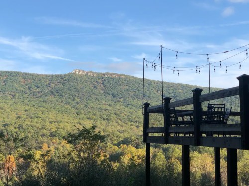A scenic view of a mountain landscape from a wooden deck with string lights and chairs, surrounded by trees.