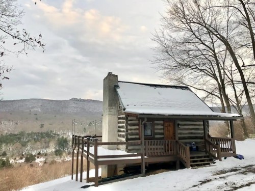 A cozy log cabin in a snowy landscape, surrounded by trees and mountains under a cloudy sky.