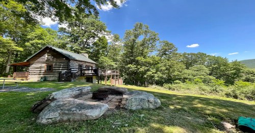 A rustic log cabin surrounded by trees, with a fire pit in the foreground and a clear blue sky above.