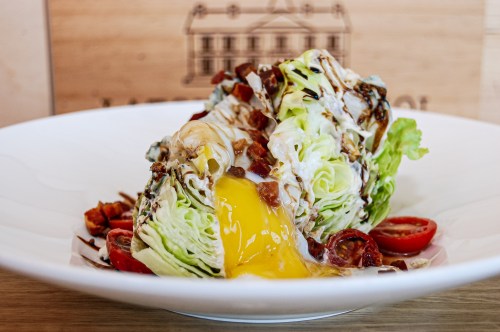 A wedge salad topped with bacon, cherry tomatoes, and a runny egg yolk, served in a white bowl.